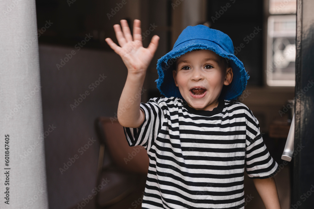 Smiling child waving hand a friendly cheerful smile to his new neighbours. Boy leaves the house go to friends and waving his hand. Boy wear blue panama hat and stripped t shirt, meeting friends. Smiling child waving hand a friendly cheerful smile to his new neighbours. Boy leaves the house go to friends and waving his hand. Boy wear blue panama hat and stripped t shirt, meeting friends.