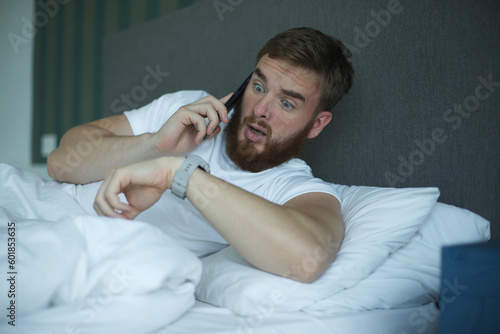 Young sleepy awaken man is just wake up with phone call, looking at his watches checking time and hurry up and yawn. Guy overslept job  photo