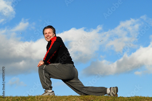 Handsome fit forties man is in grass field performing some stretching exercises with a beautiful smile on his face.There is copyspace on the sky backround.