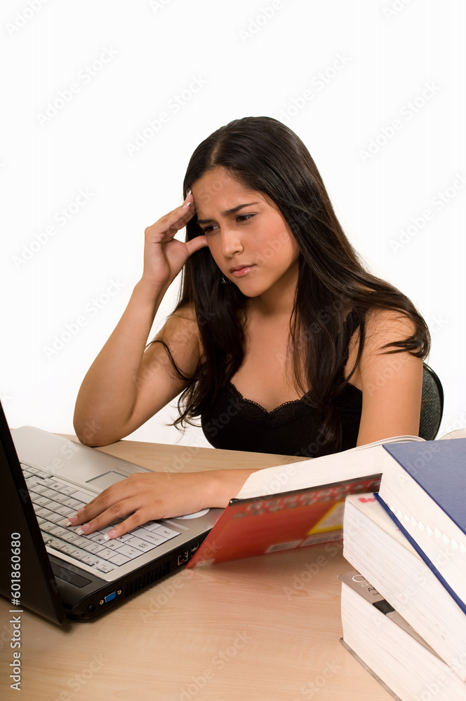 Young Hispanic woman sitting in front of desk reading a textbook and typing on a laptop with pained expression Young Hispanic woman sitting in front of desk reading a textbook and typing on a laptop with pained expression