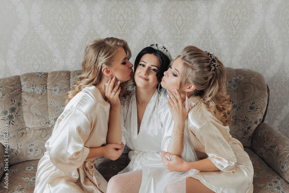 A brunette bride and her blonde friends are sitting on the sofa in the bride's room, wearing bathrobes, smiling and posing. Wedding portrait wedding photo in a light key. Morning of the bride. A brunette bride and her blonde friends are sitting on the sofa in the bride's room, wearing bathrobes, smiling and posing. Wedding portrait wedding photo in a light key. Morning of the bride.