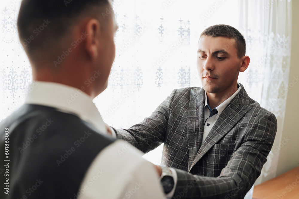 Fototapeta premium Photo of two men in classic suits. A handsome young man mends the shirt of another man standing in front of him. Business style. Stylish men. Clock