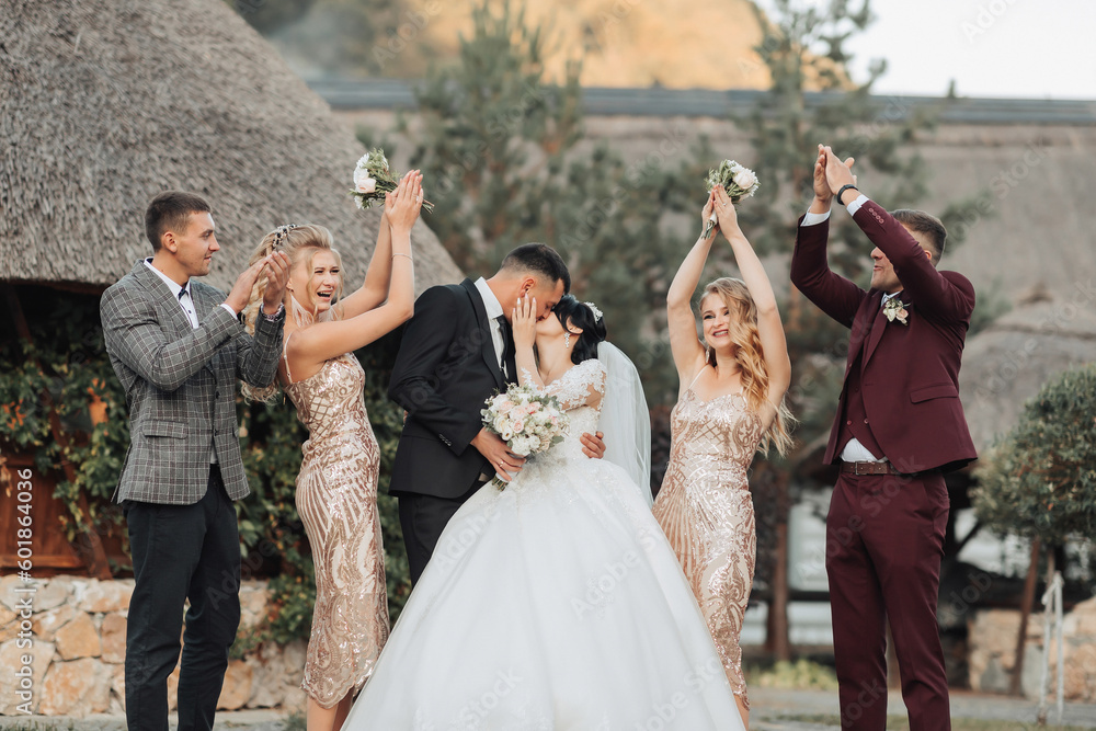 Fototapeta premium Wedding portrait. Front view of newlyweds standing and kissing, between friends, friends having fun. Girls in golden dresses hold bouquets, the bride in a voluminous dress, the groom