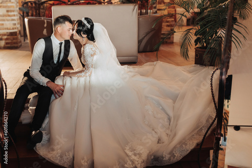Wedding portrait of the bride and groom. Indoor photography with beautiful light. Stylish Groom in a black vest kisses a brunette bride on the cheek, sitting hugging indoors. Couple in love