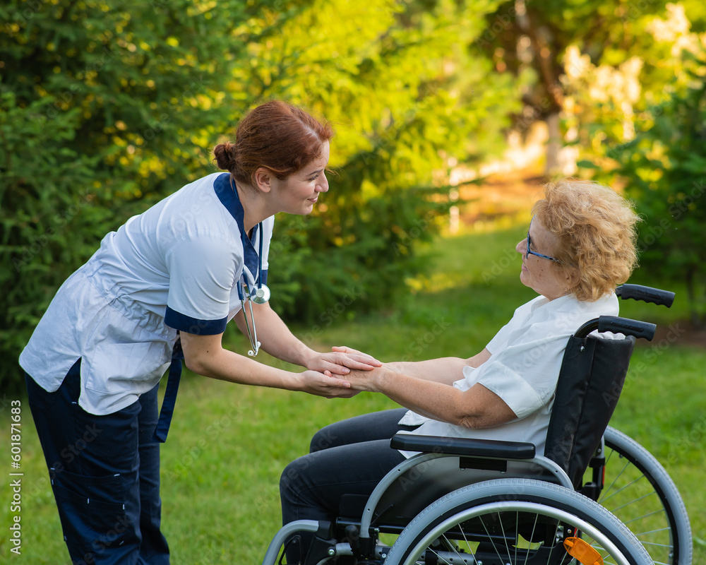 A nurse holds an elderly caucasian woman in a wheelchair by the hand as support. Nurse walks with a patient in the park. A nurse holds an elderly caucasian woman in a wheelchair by the hand as support. Nurse walks with a patient in the park.