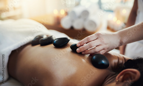 Woman, hands and rocks in back massage at spa for skincare, beauty or relaxation on bed indoors. Hand of masseuse applying hot rock or stones on female for physical therapy or treatment at resort