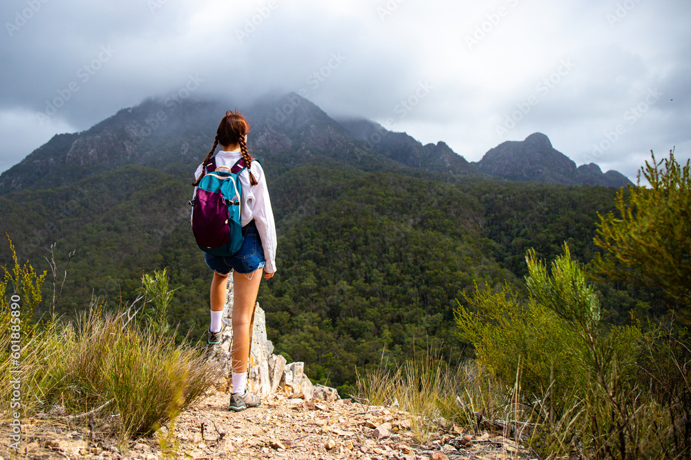 Naklejka premium beautiful and brave girl standing at the top of the hill with large mountain in front of her; mighty mount barney in queensland, australia seen from yellow pinch lookout