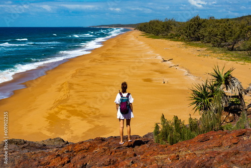 backpacker girl in white dress standing on the cliff admiring the panorama of beautiful beach with orange sand; pacific coast of australia in deepwater national park, queensland photo