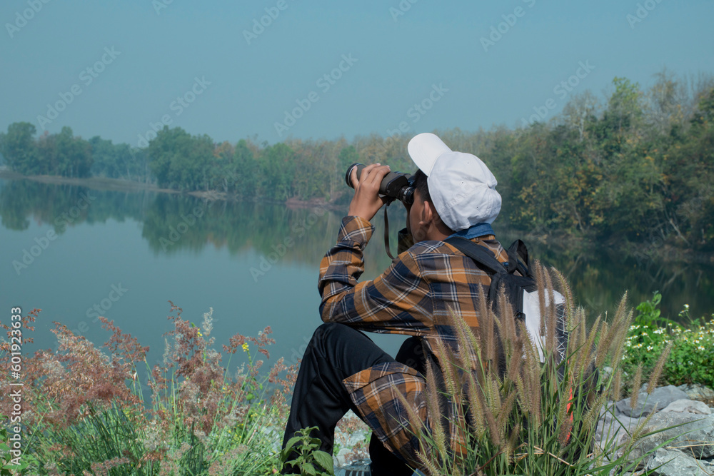 Asian boy in plaid shirt wears white cap, holding binoculars, standing on reservoir ridge during summer vacation and birdwatching activity, soft and selective focus, nature study and hobby concept.
