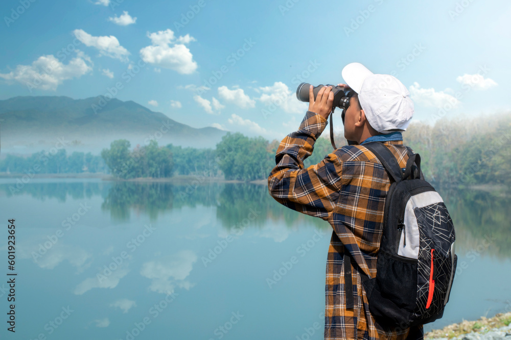 Obraz premium Asian boy in plaid shirt wears white cap, holding binoculars, standing on reservoir ridge during summer vacation and birdwatching activity, soft and selective focus, nature study and hobby concept.