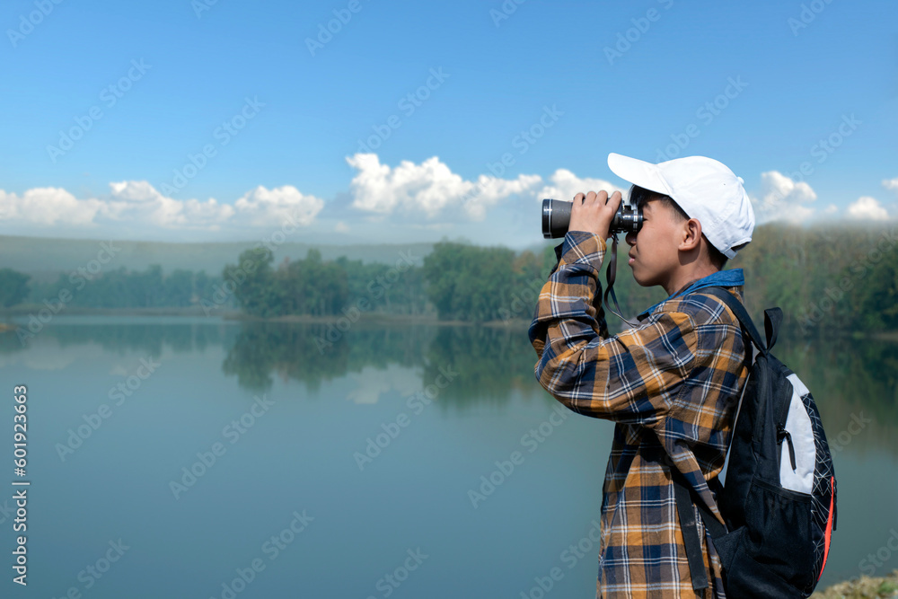 Fototapeta premium Asian boy in plaid shirt wears white cap, holding binoculars, standing on reservoir ridge during summer vacation and birdwatching activity, soft and selective focus, nature study and hobby concept.