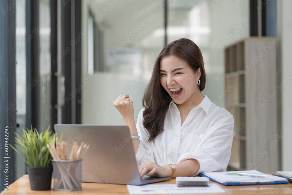 An Asian businesswoman celebrating victory success, an employee inside the office reading good news, using a laptop at work inside the office holding hand up, and a happy triumph gesture.