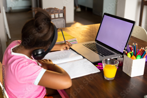 African american girl doing schoolwork using headphones and laptop at home, copy space on screen