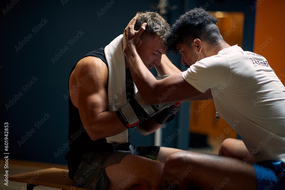 Coach helping young boxing player mentally prepare, holding his head and talking to him, in dressing room sitting on bench.