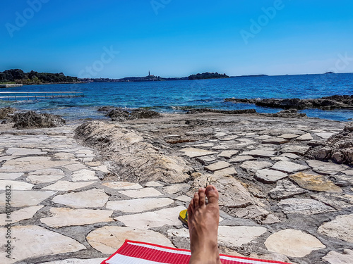 Relaxing on the beach. A human foot lying on a stony beach on a red towel. In the back there is bigger city with tall church tower. Clear and sunny day, perfect for suntanning. photo