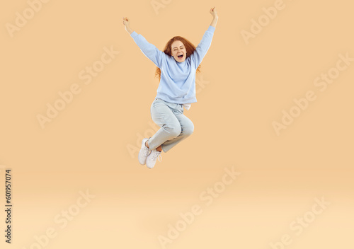 Overjoyed excited teenage girl who feels euphoria joyfully shouts and jumps high with raised arms. Red-haired curly Caucasian girl in casual outfit jumping isolated on light beige background.