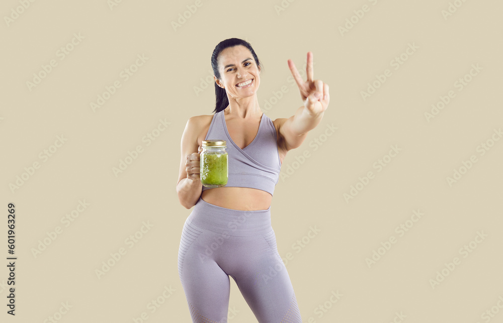 Attractive Caucasian woman holding jar of beneficial fruit smoothie and smiling showing victory gesture calling for healthy lifestyle or diet dressed in fitness clothes standing in studio Attractive Caucasian woman holding jar of beneficial fruit smoothie and smiling showing victory gesture calling for healthy lifestyle or diet dressed in fitness clothes standing in studio