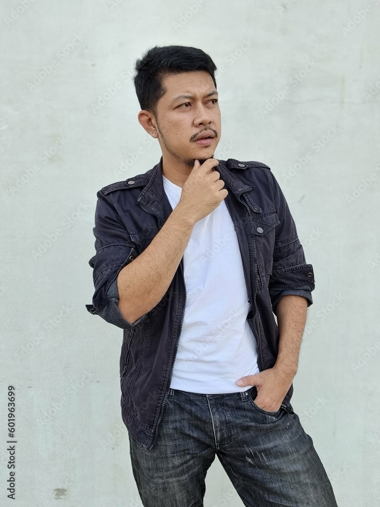 Cool modern young hipster man with a stylish hairstyle in a white T-shirt puts on a vintage black denim jacket. Attractive urban guy posing near a white wall. Fashionable spring menswear.