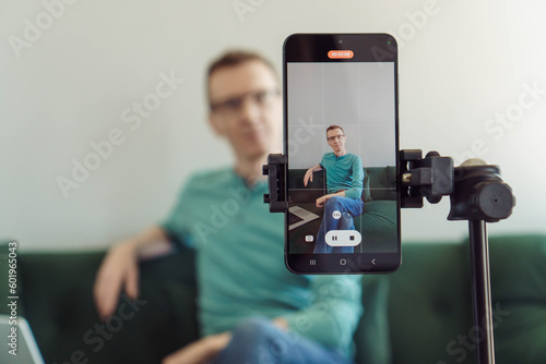 Unfocused man is sitting on sofa in background, focusing on phone on tripod with picture of camera shooting. Blogger. Modern technologies. Online training. Video recording of trainings. Video chat.