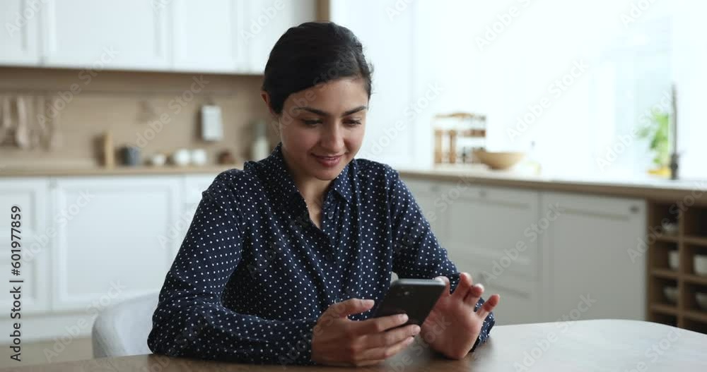 Smiling Indian woman holds cellphone sit in kitchen, read message, use new mobile application, enjoy e-services to easier life, feel satisfied use modern phone, share sms enjoy remote communication Smiling Indian woman holds cellphone sit in kitchen, read message, use new mobile application, enjoy e-services to easier life, feel satisfied use modern phone, share sms enjoy remote communication