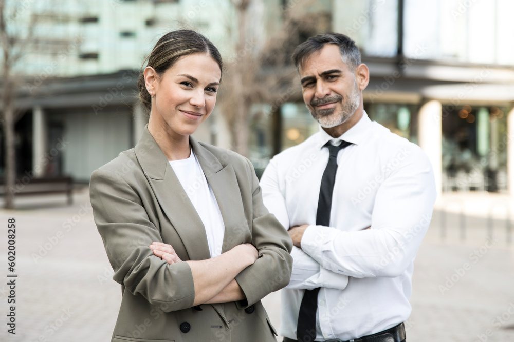 Confident and smiling business people looking at camera in the street with their arms crossed. Young businesswoman with a senior businessman behind looking satisfied. Confident and smiling business people looking at camera in the street with their arms crossed. Young businesswoman with a senior businessman behind looking satisfied.