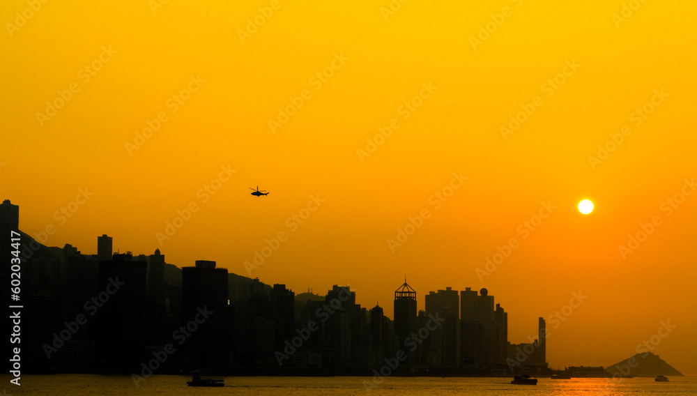 Golden hour sunset over silhouette of Hong Kong skyline with helicopter flying and boats on Victoria Harbour