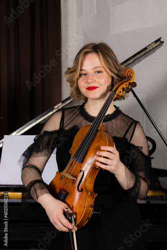 Young elegant blonde girl posing with a violin. Portrait of a female violinist playing, performing against a piano.