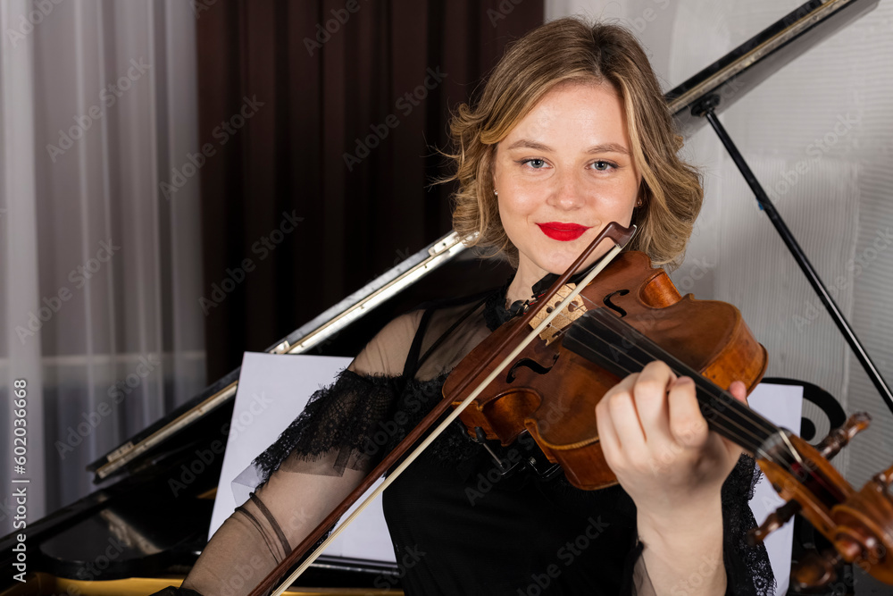 Young elegant blonde girl posing with a violin. Portrait of a female violinist playing, performing against a piano.