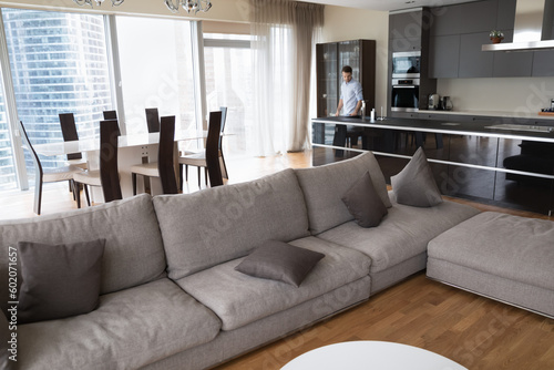 Elegant interior of modern urban apartment with large window and city skyscraper view. Hotel senior suite room, studio with big comfortable couch and young man in kitchen in background