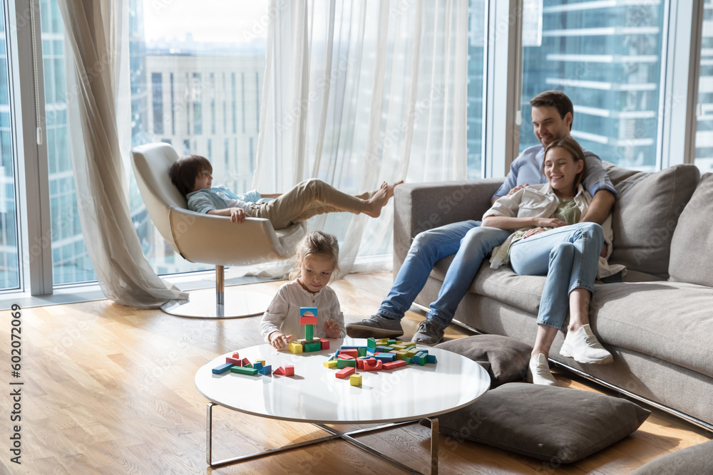 Fototapeta premium Couple of happy young parents resting on sofa in city apartment with large window, watching children, Two kids enjoying leisure at home, constructing toy building blocks, relaxing in armchair