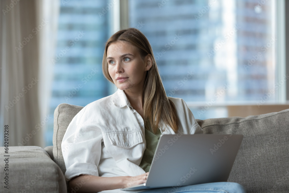 Dreamy beautiful young freelancer woman working at laptop, sitting on home couch, using job professional application on computer, looking away, thinking on wireless internet technology Dreamy beautiful young freelancer woman working at laptop, sitting on home couch, using job professional application on computer, looking away, thinking on wireless internet technology