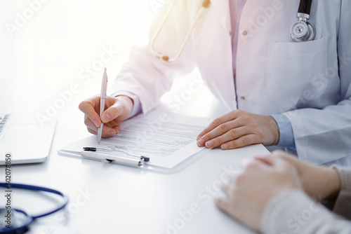 Doctor and patient sitting at the table in clinic. The focus is on female physician's hands filling up the medication history record form or checklist, close up. Medicine concept