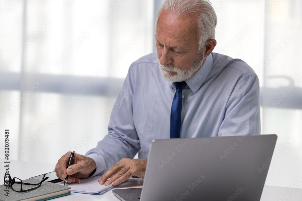 Confident senior businessman working and taking notes in his modern office. Mature CEO or director using laptop and signing documents Confident senior businessman working and taking notes in his modern office. Mature CEO or director using laptop and signing documents