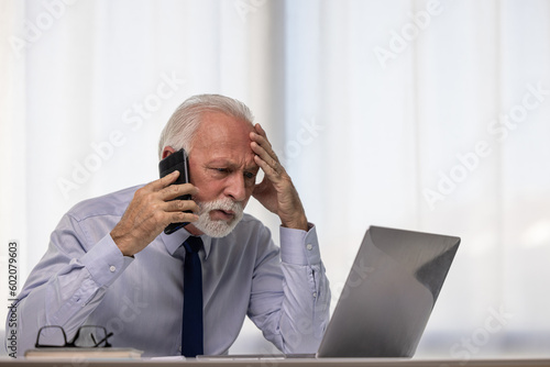 Shocked senior businessman talking on smartphone and using laptop, hearing bad news, unexpected debt or bankruptcy, financial problems. Frustrated upset CEO making phone call