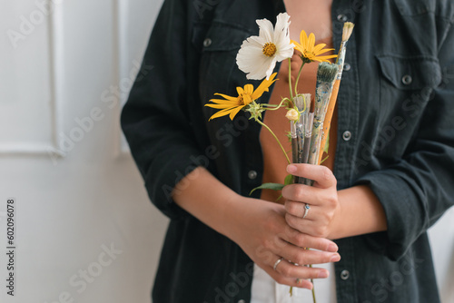 Close-up of young artist holding bouquet made of flowers and paint brushes in studio