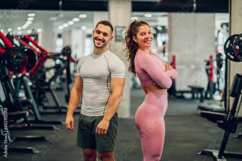 The sporty happy muscular couple is posing in a gym while smiling at the camera.