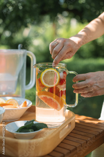 Woman preparing a jug of water infused with assorted fresh fruit and mint leaves in the garden