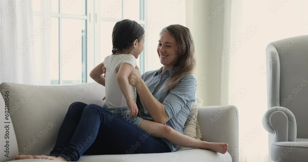 Cheerful mom cuddling cute girl on lap, talking to child with love, tenderness, care, laughing, smiling, resting on home sofa. Sweet kid speaking to mother, showing, pointing hand away Cheerful mom cuddling cute girl on lap, talking to child with love, tenderness, care, laughing, smiling, resting on home sofa. Sweet kid speaking to mother, showing, pointing hand away