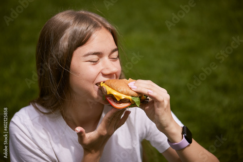 a young girl takes out a burger from a paper bag sitting on the green grass  the concept of food delivery  street food