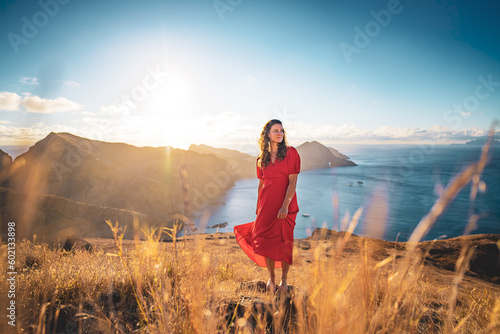 Woman in red dress on yellow field enjoys the morning atmosphere from the foothills of a volcanic island in the Atlantic Ocean. São Lourenço, Madeira Island, Portugal, Europe.