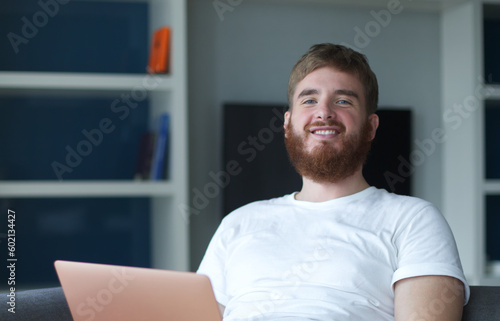 Young happy positive man use laptop computer at home in living room sit on couch looking at camera and smile. Freelancer guy, freelance distant job