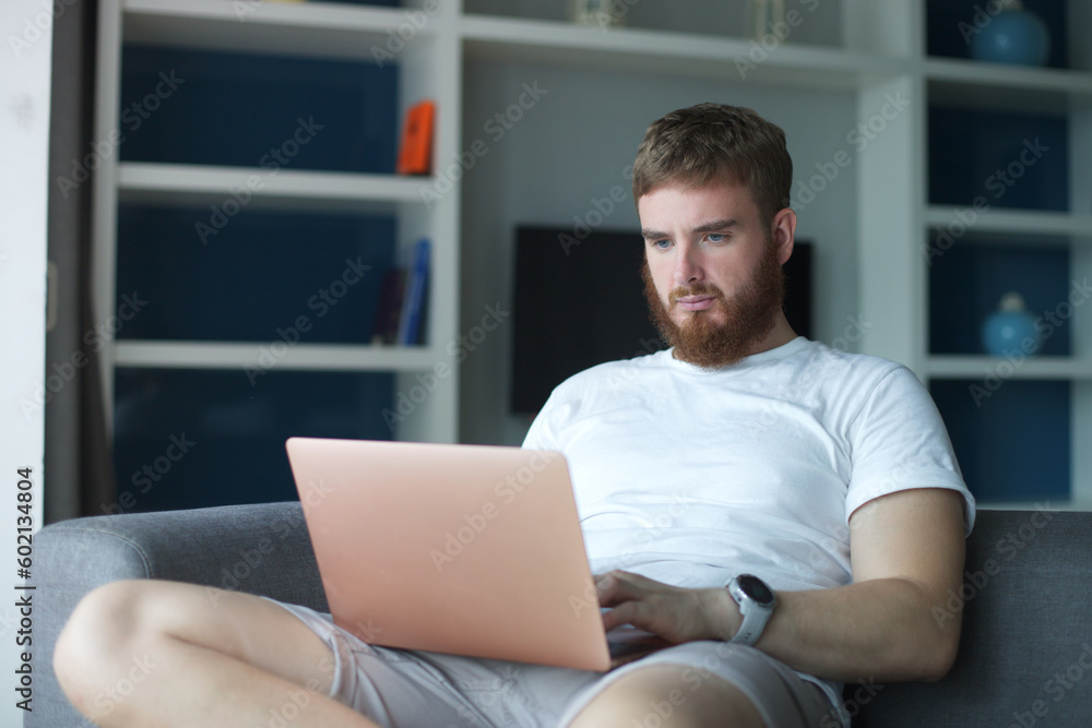 Young happy positive man use laptop computer at home in living room sit on couch looking at camera and smile. Freelancer guy, freelance distant job Young happy positive man use laptop computer at home in living room sit on couch looking at camera and smile. Freelancer guy, freelance distant job