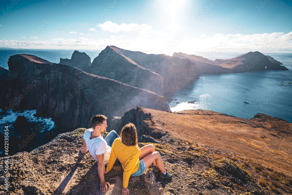Back view of a seated young tourist couple enjoying the coastal landscape of Madeira Island in the Atlantic Ocean in the morning. São Lourenço, Madeira Island, Portugal, Europe.