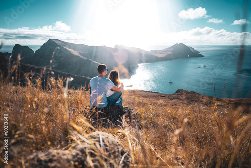 Back view of a tourist couple sitting on a hill, overlooking the coastal landscape of Madeira Island in the Atlantic Ocean in the morning. São Lourenço, Madeira Island, Portugal, Europe.