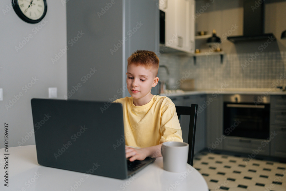 Portrait of adorable redhead child boy with freckles looking to laptop screen typing doing homework. Cute preteen boy surfing internet on notebook computer sitting at table with coffee cup at home. Portrait of adorable redhead child boy with freckles looking to laptop screen typing doing homework. Cute preteen boy surfing internet on notebook computer sitting at table with coffee cup at home.