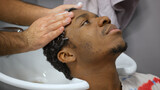 African american man client in barbershop. Hands of barber are washing his hair with shampoo over the sink, side view. Hairstylist hairdresser is taking care man's hairs in beauty haircut salon.