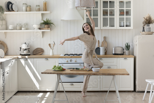 Cheerful excited active young woman having fun in home kitchen  dancing at table with organic food  fresh vegetables for salad  celebrating culinary success  feeling full of energy due to healthy diet