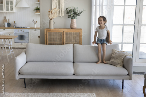 Cheerful excited little kid jumping on couch at home, standing on sofa, laughing, smiling, playing active games, enjoying activities, leisure, playtime in home interior, having fun