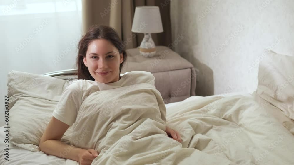 An attractive young woman is sitting on a bed in the early sunny morning, smiling and looking at the camera An attractive young woman is sitting on a bed in the early sunny morning, smiling and looking at the camera