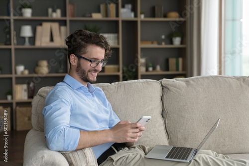 Happy positive young freelance entrepreneur man enjoying Internet technology at home, using mobile phone, laptop for work communication, resting on sofa with digital gadgets, looking at screen